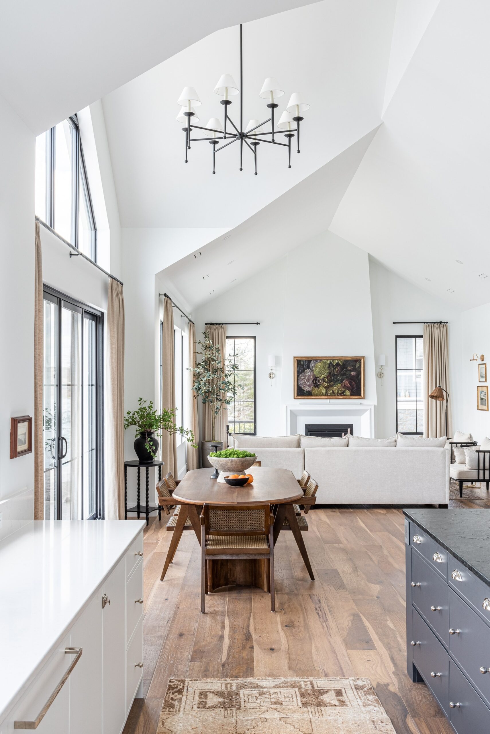 Looking from the kitchen to the casual dining and main living area, featuring Hudson Valley Lighting's London Chandelier. The black finish with white shades draws your eye upward to the high vaulted ceilings and abundant natural light